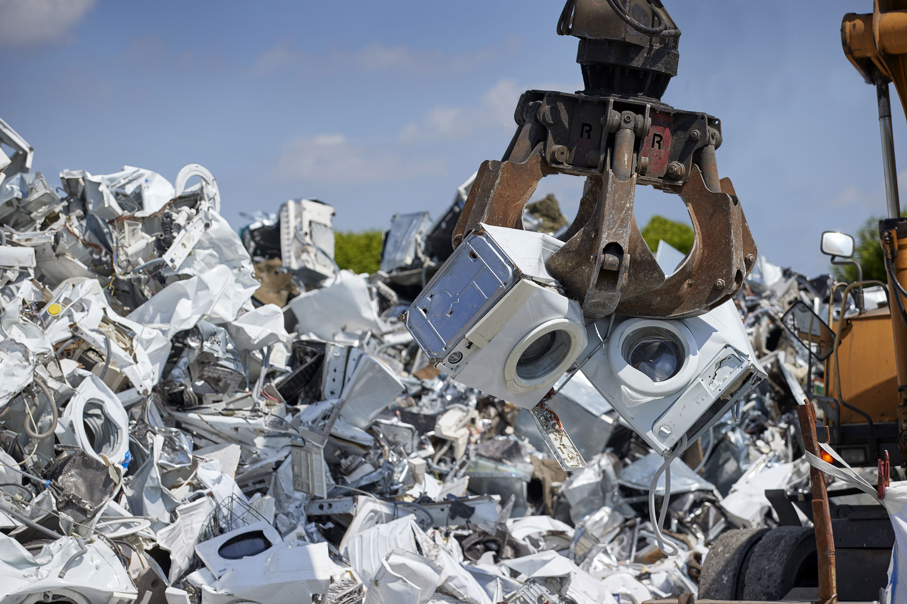 A claw crane lifts two scrap washing machines from a large pile of electronic waste in a scrap yard.