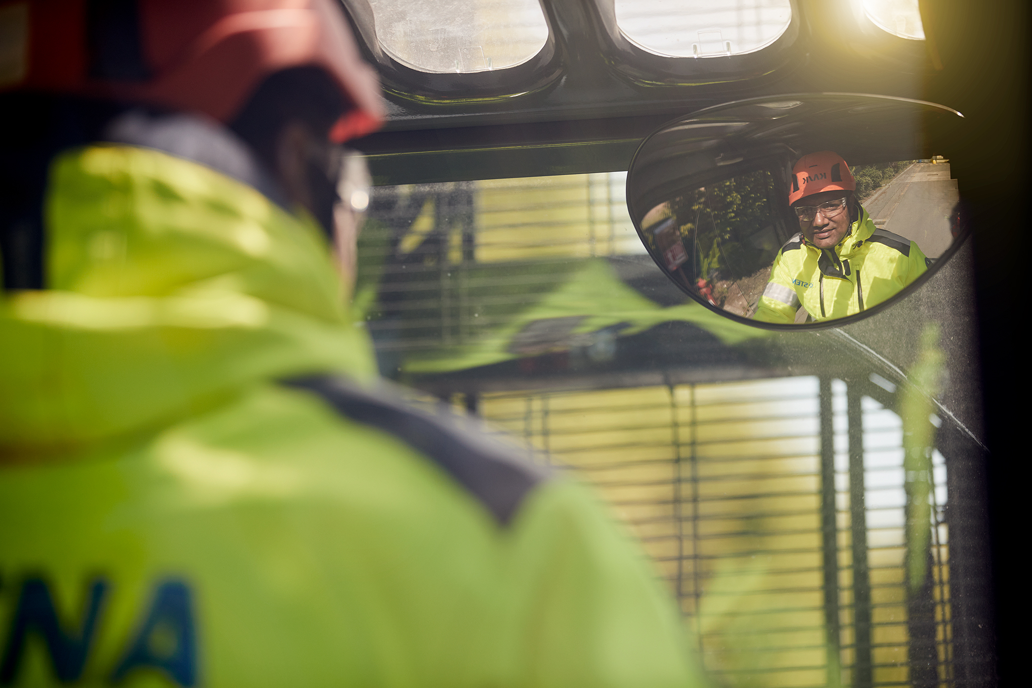 Arbejder i synlig jakke og sikkerhedshjelm, reflekteret i et køretøjsspejl.