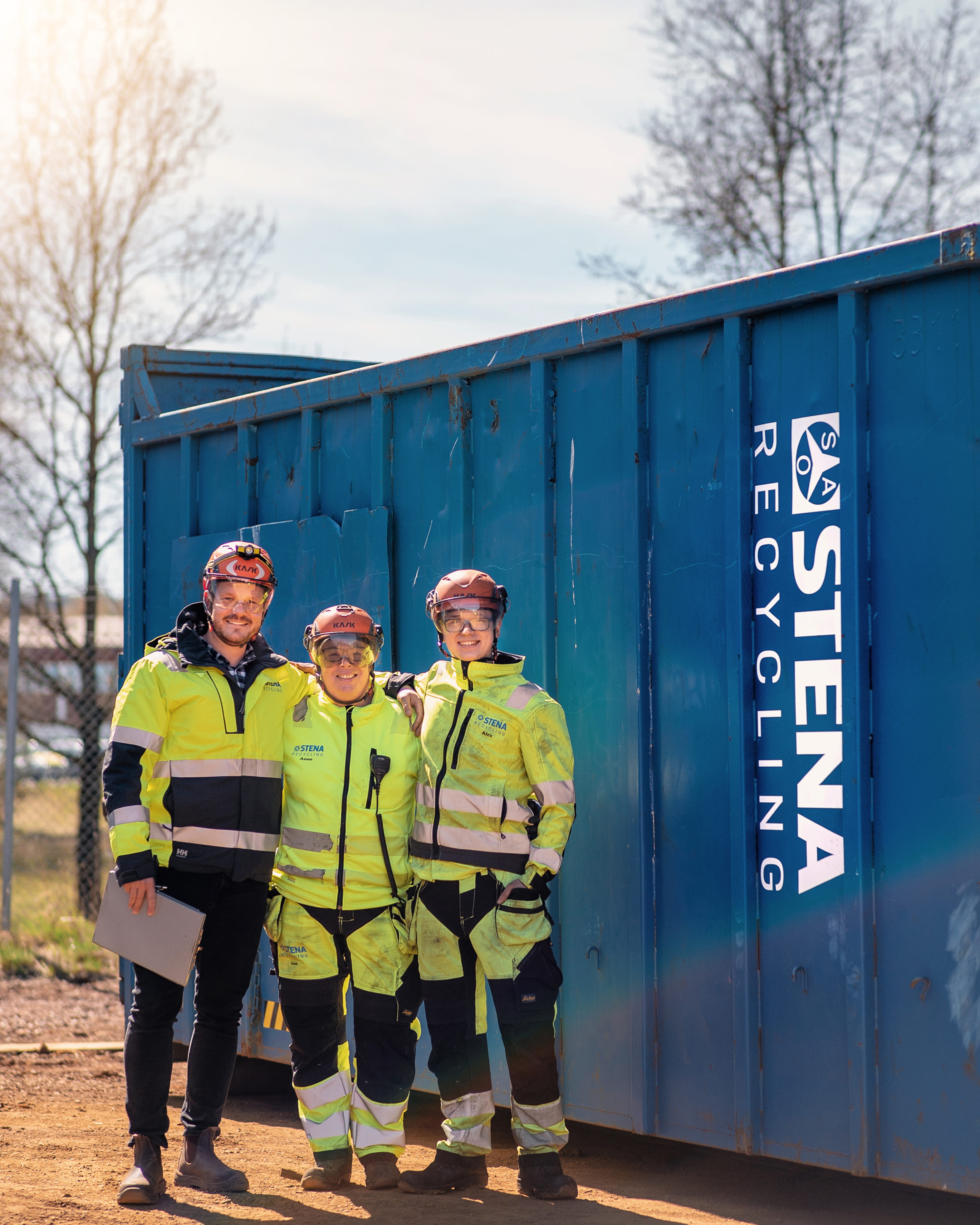 Tre Stena Recycling-eksperter klædt i reflekterende arbejdstøj, beskyttelseshjelm og beskyttelsesbriller, står foran en blå Stena Recycling-stålcontainer, holder hinanden om skuldrene og smiler til fotografen.