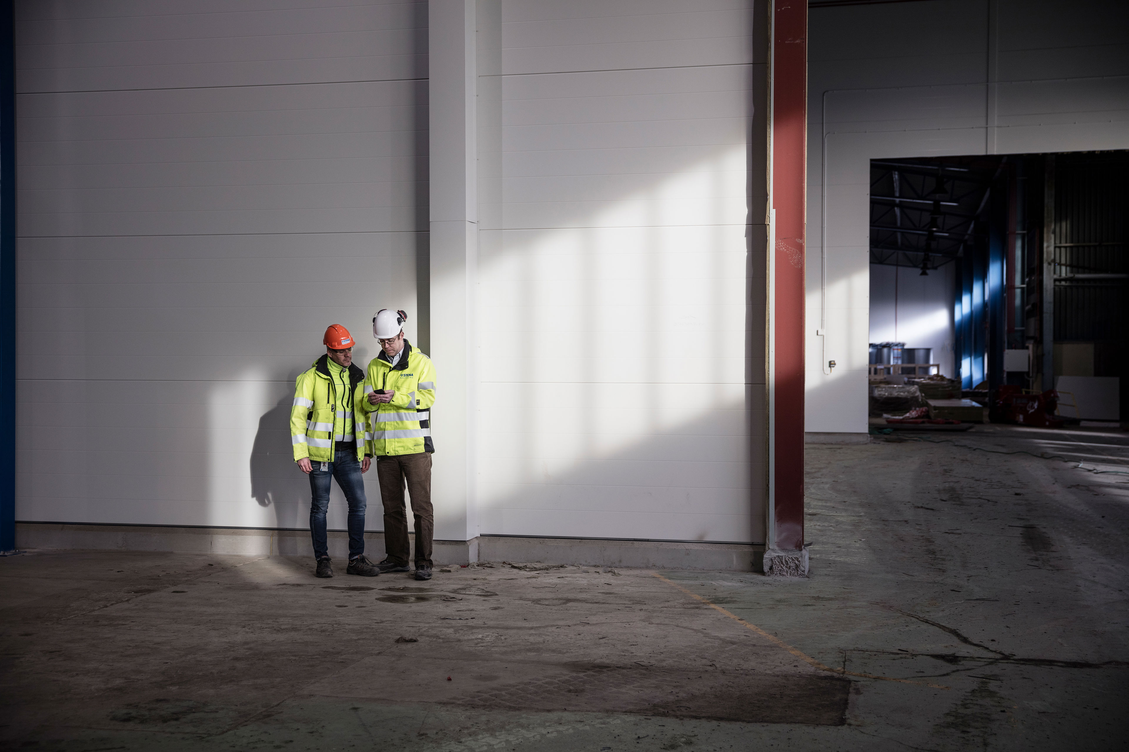 Two male Stena Recycling employees in protective gear look at a mobile phone together in a recycling facility.
