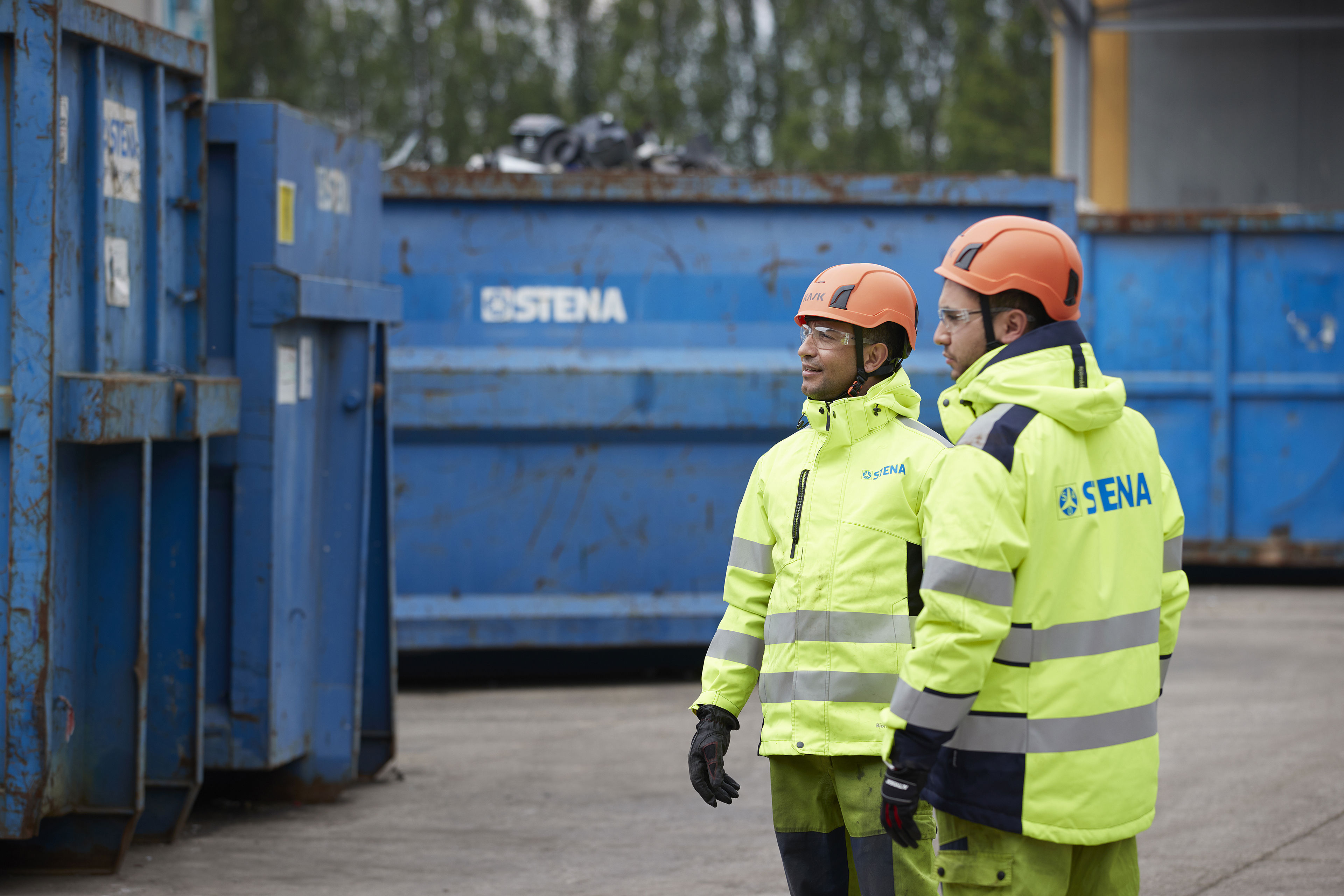 Two recycling experts dressed in hardhats and reflective Stena Recycling work clothes standing  beside a couple of large blue recycling containers in a recycling yard.