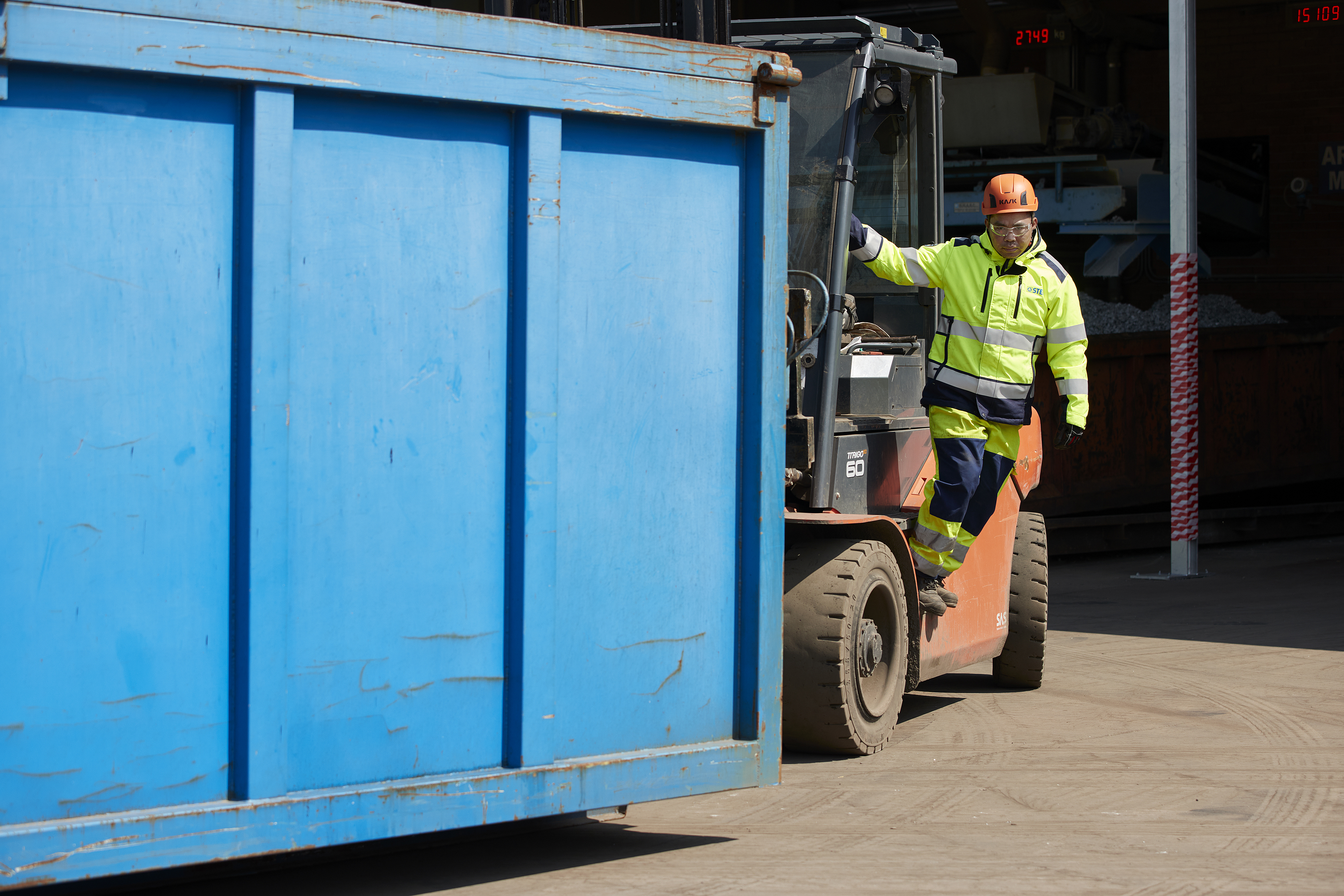 A male Stena Recycling employee in hi-vis protective gear climbs out of a forklift truck next to a scrap collection container.