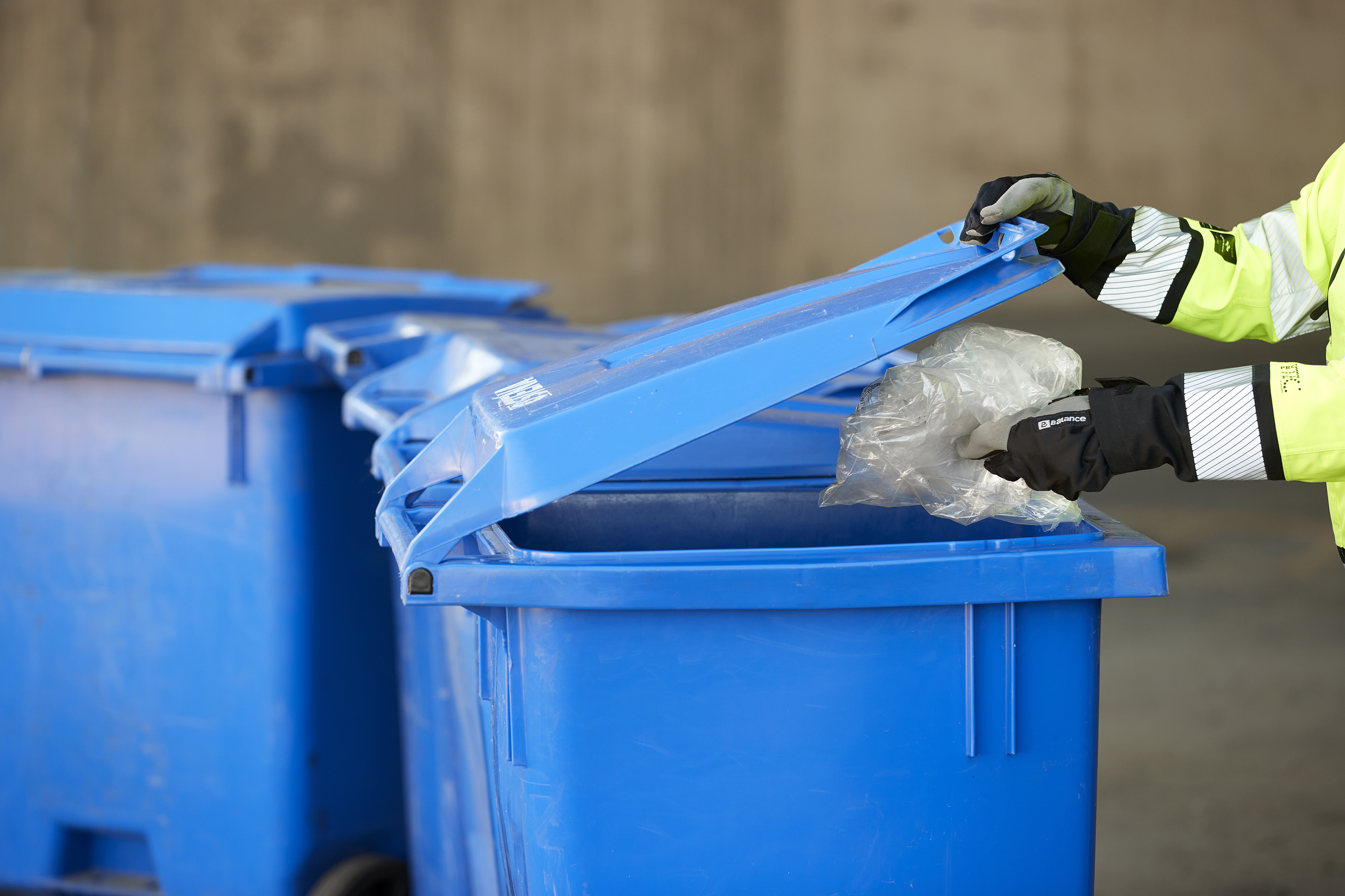 Hands wearing work gloves opening the lid of a blue plastic recycling bin.
