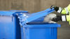 Hænder iført arbejdshandsker åbner låget på en blå plastikbeholder til genbrug.