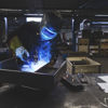 A worker in protective gear welds a piece of metal in a factory environment.
