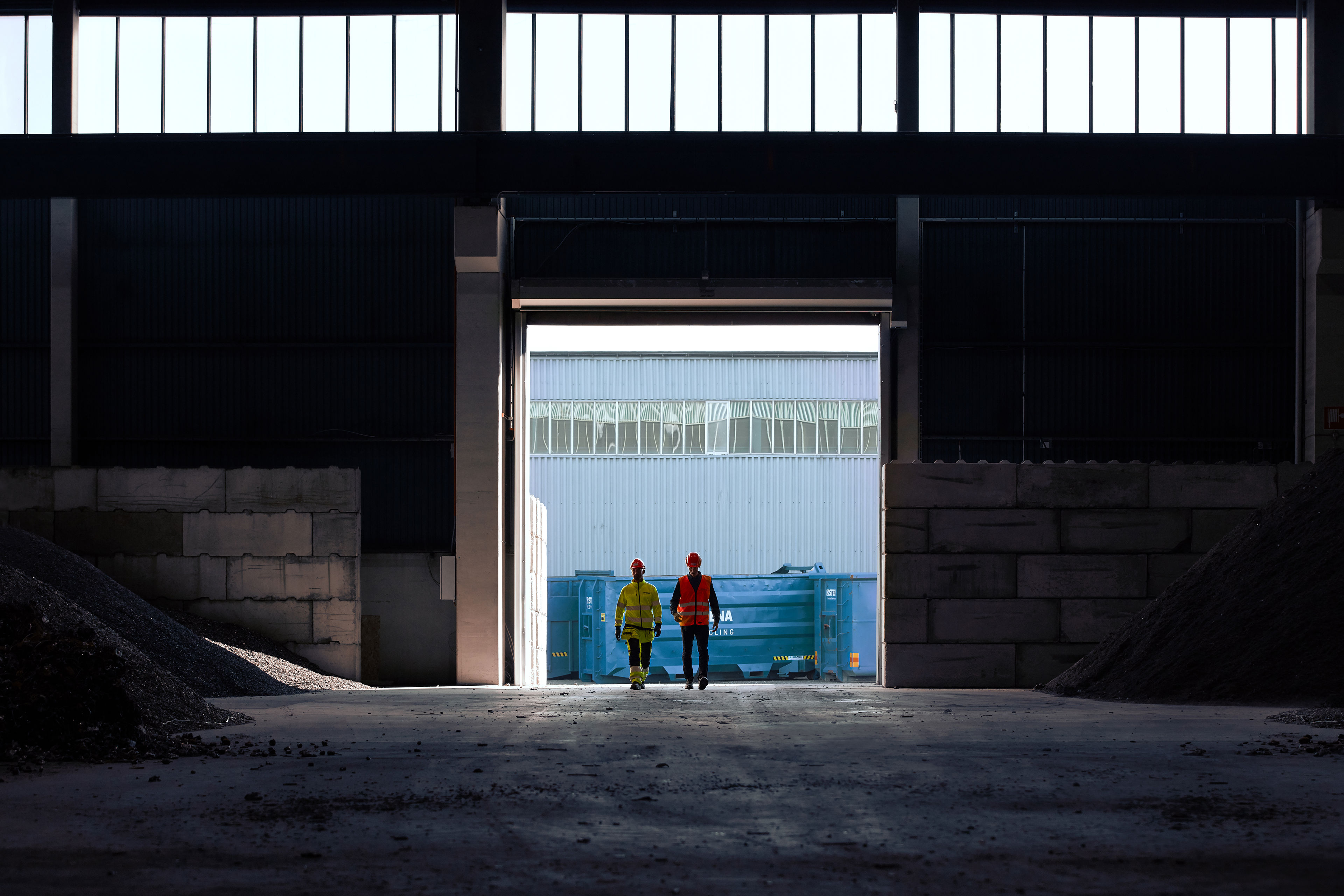 Two recycling experts dressed in hardhats and reflective work clothes entering a warehouse containing heaps of recycled material.